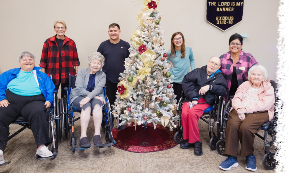 Group posing around a decorated Christmas tree in a room with a sign on the wall reading, 
