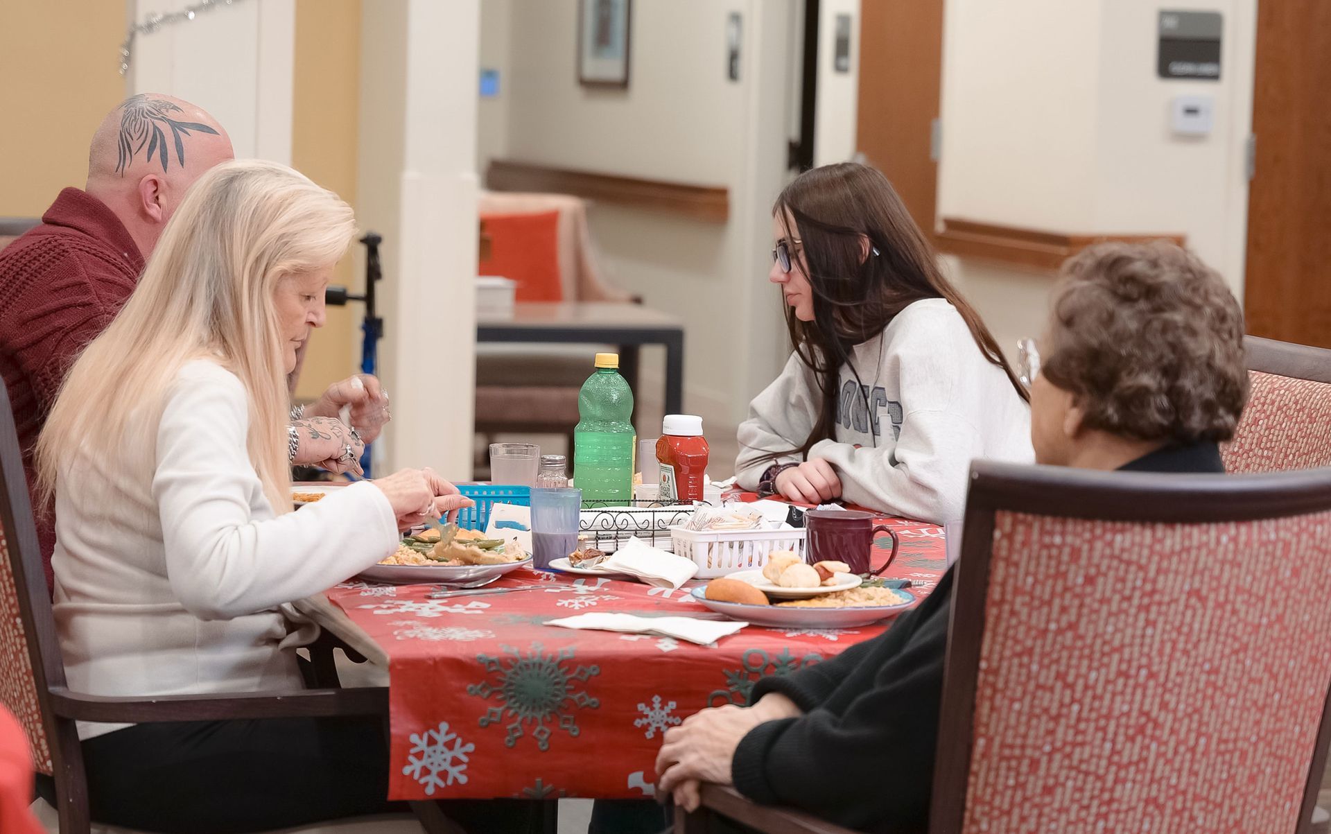Four people sit around a table with a red holiday-themed tablecloth, eating a meal in a brightly lit indoor space.