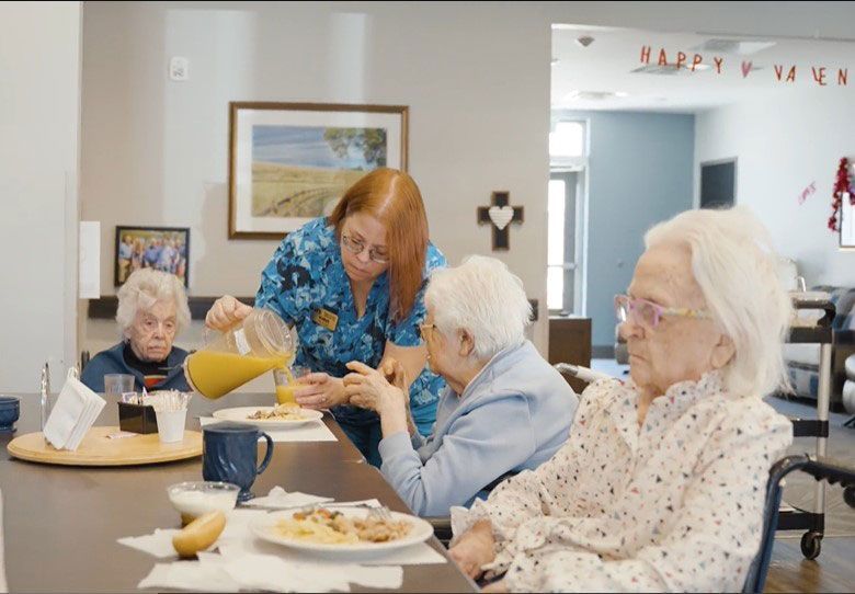 A staff member in a blue patterned shirt pours orange juice for a person at a dining table in a senior living facility.