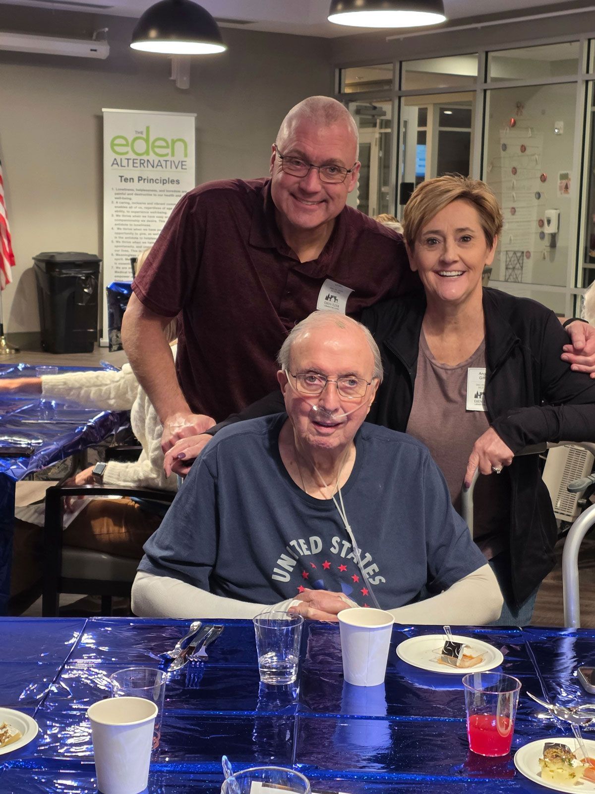 Three people smile together behind a blue-covered dining table at an indoor event, with a branded sign in the background.