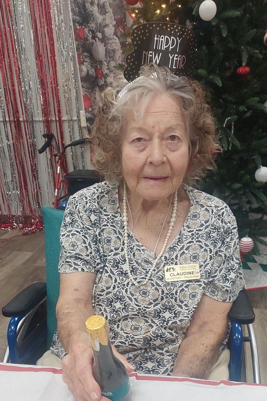 An elderly person sitting in a wheelchair holding a bottle, wearing a 'Happy New Year' headband in a decorated room.