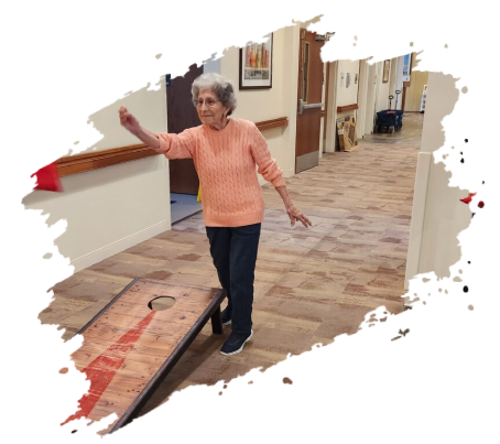 Elderly woman throwing a bean bag at a cornhole board in a hallway.