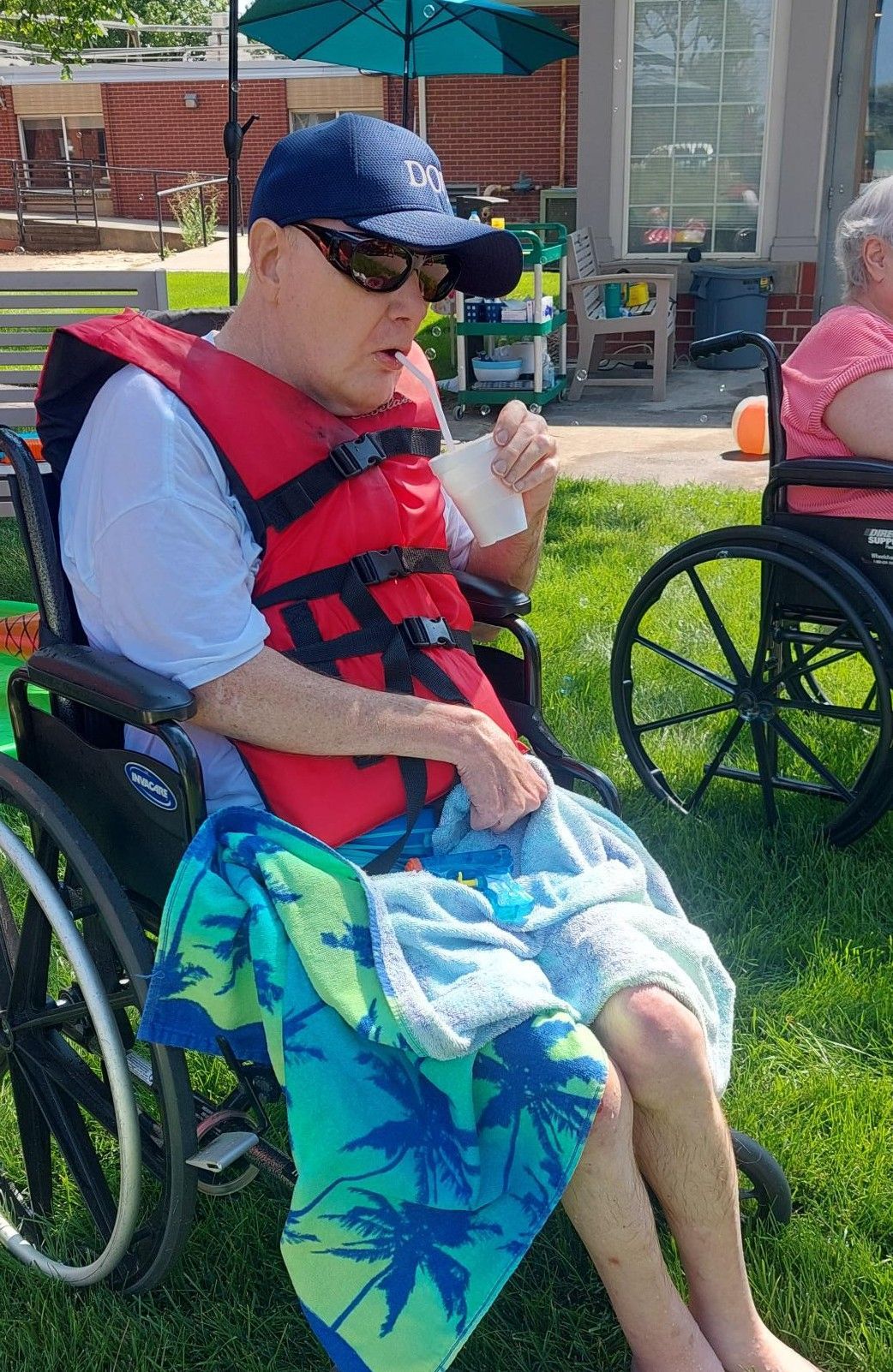 A person in a wheelchair, wearing a red life vest, hat, and sunglasses, sits on a grassy lawn with a towel on their lap.
