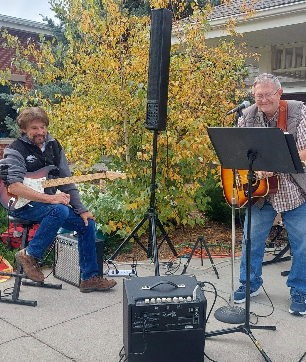Two musicians play guitars and perform outdoors on a patio with autumn foliage in the background.