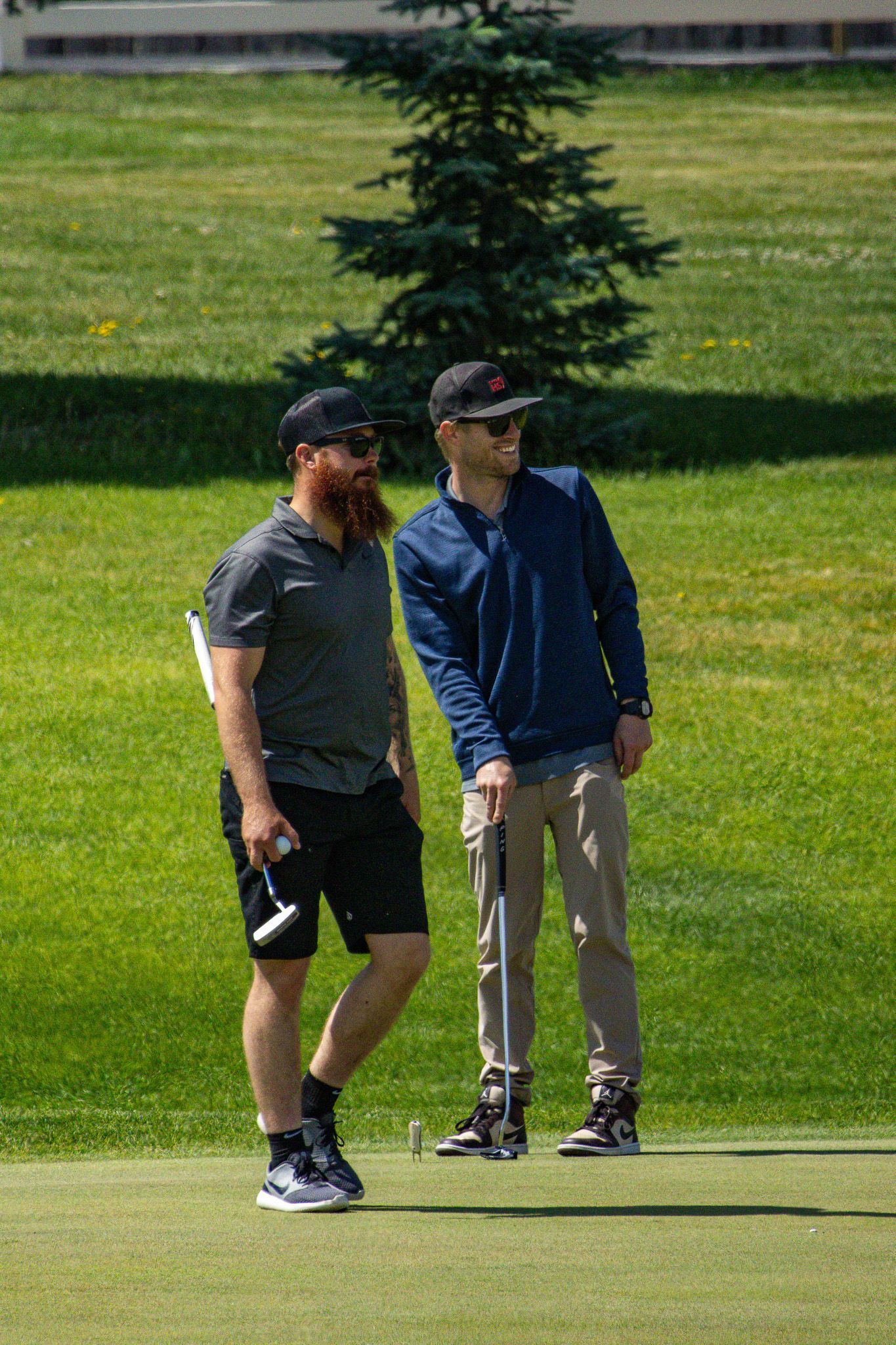 Two men standing on a grassy golf course, one holding a phone, with a small tree behind them.