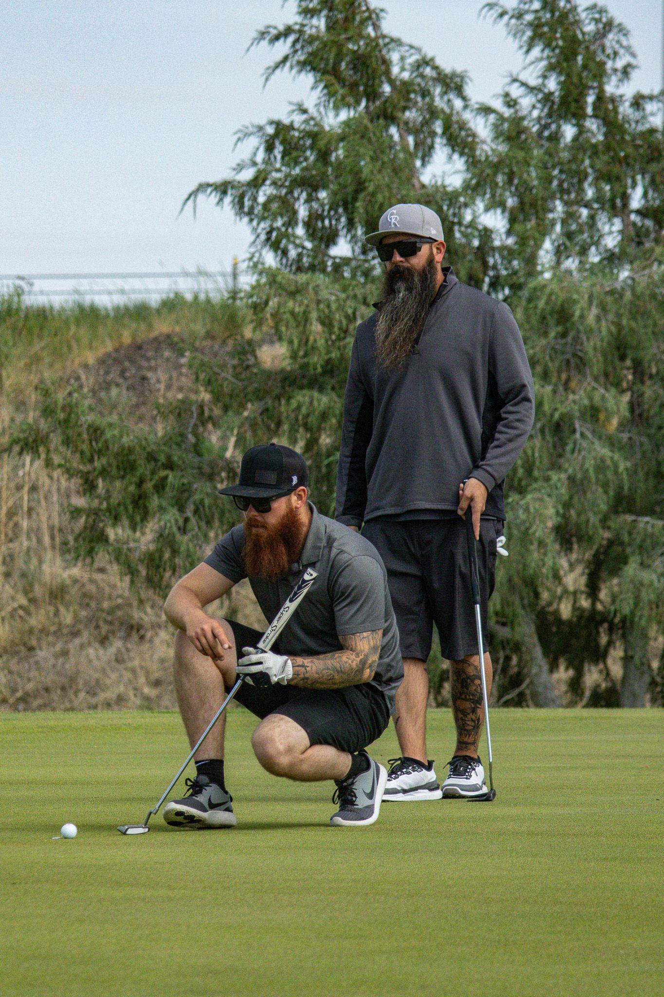 Two golfers on a green, one crouching to line up a putt while the other stands nearby holding a club.
