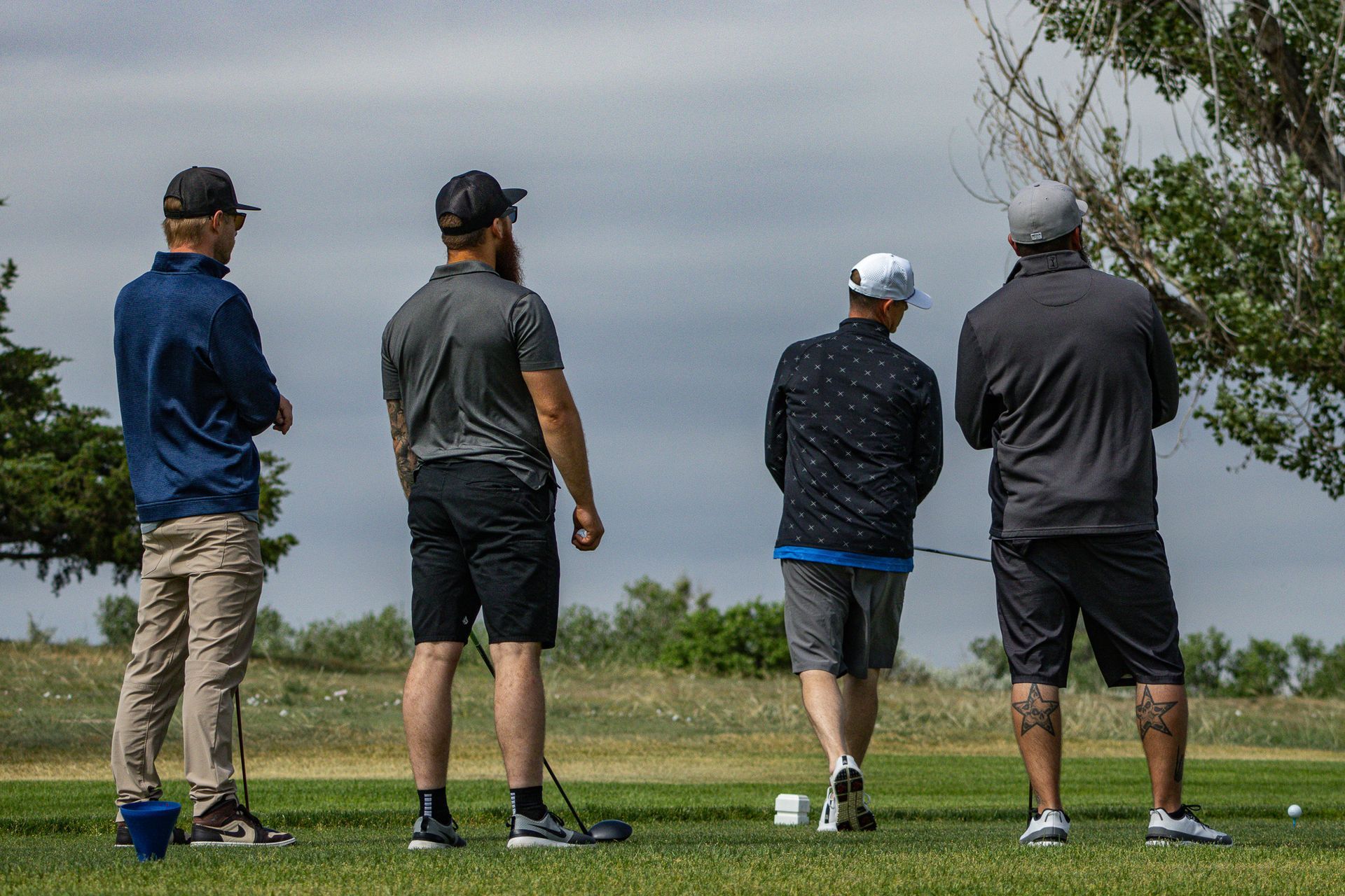 Four golfers standing on a course, viewed from behind, waiting near a tee in cloudy weather