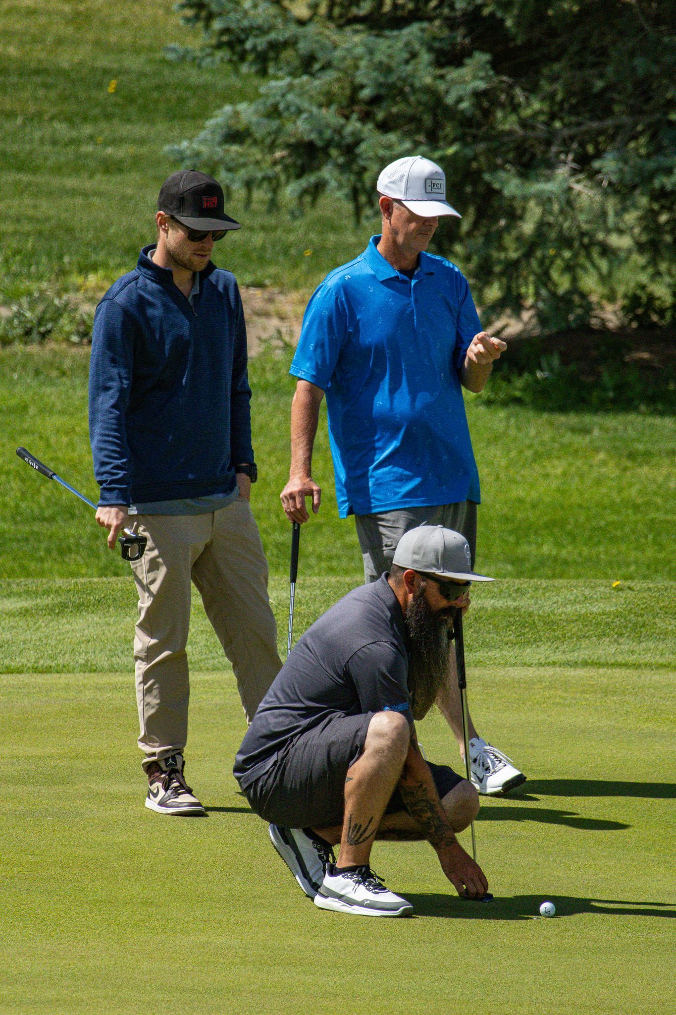 Three golfers on a green, one crouching to line up a putt while two others watch.