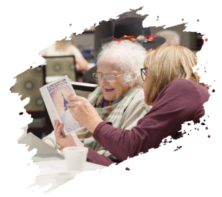 Woman shows a brochure to an elderly woman, both smiling indoors.