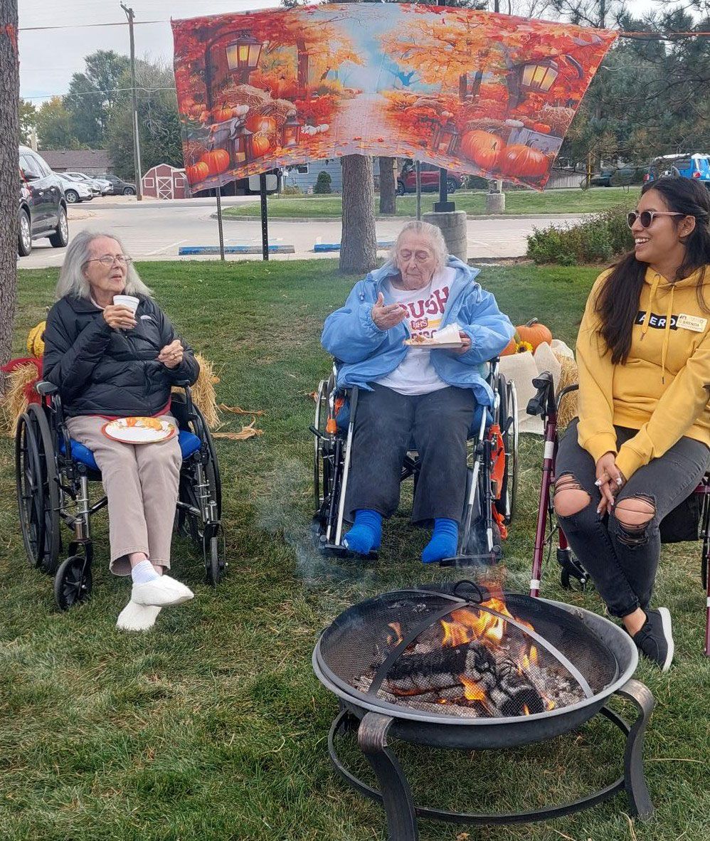 Two people in wheelchairs and one person sitting in a lawn chair around a small outdoor fire pit on a grassy lawn.