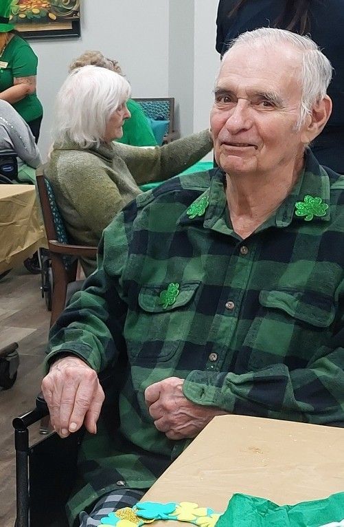 An older person in a green and black plaid shirt decorated with shamrocks sits at a table in a bright, decorated room.