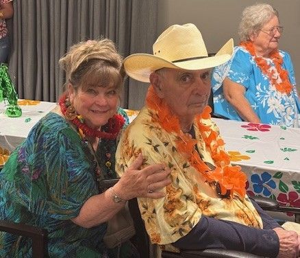 A smiling person in a teal tropical shirt sits beside another in a cowboy hat and floral shirt with an orange lei.