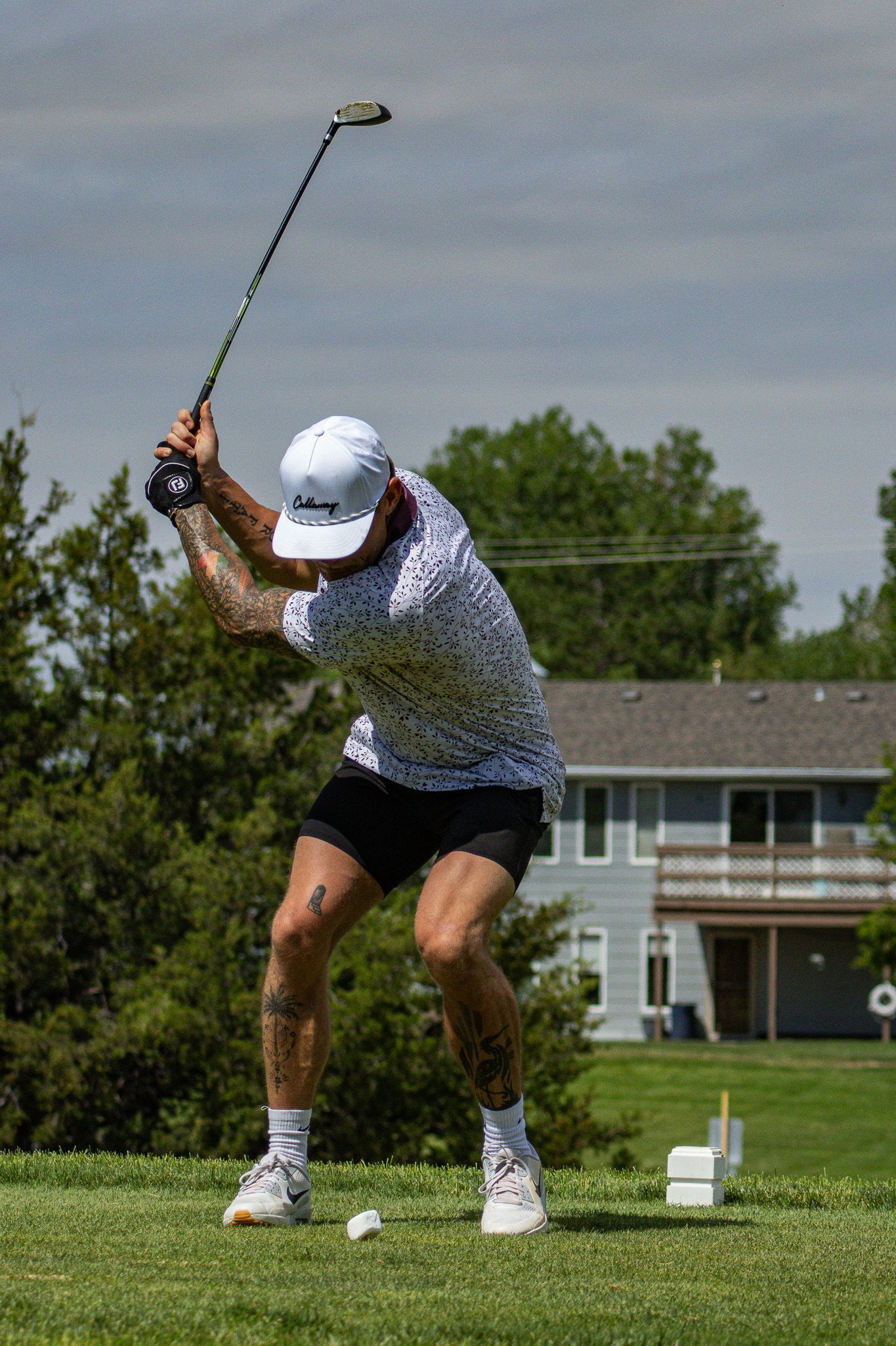 Golfer mid-swing on a grassy course, with a tee and suburban houses in the background
