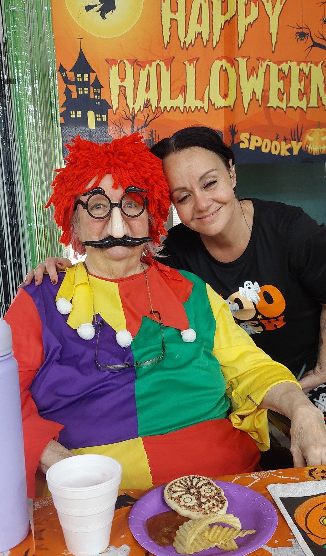 A person in a colorful jester costume and red wig sits with another person smiling in front of a Happy Halloween banner.