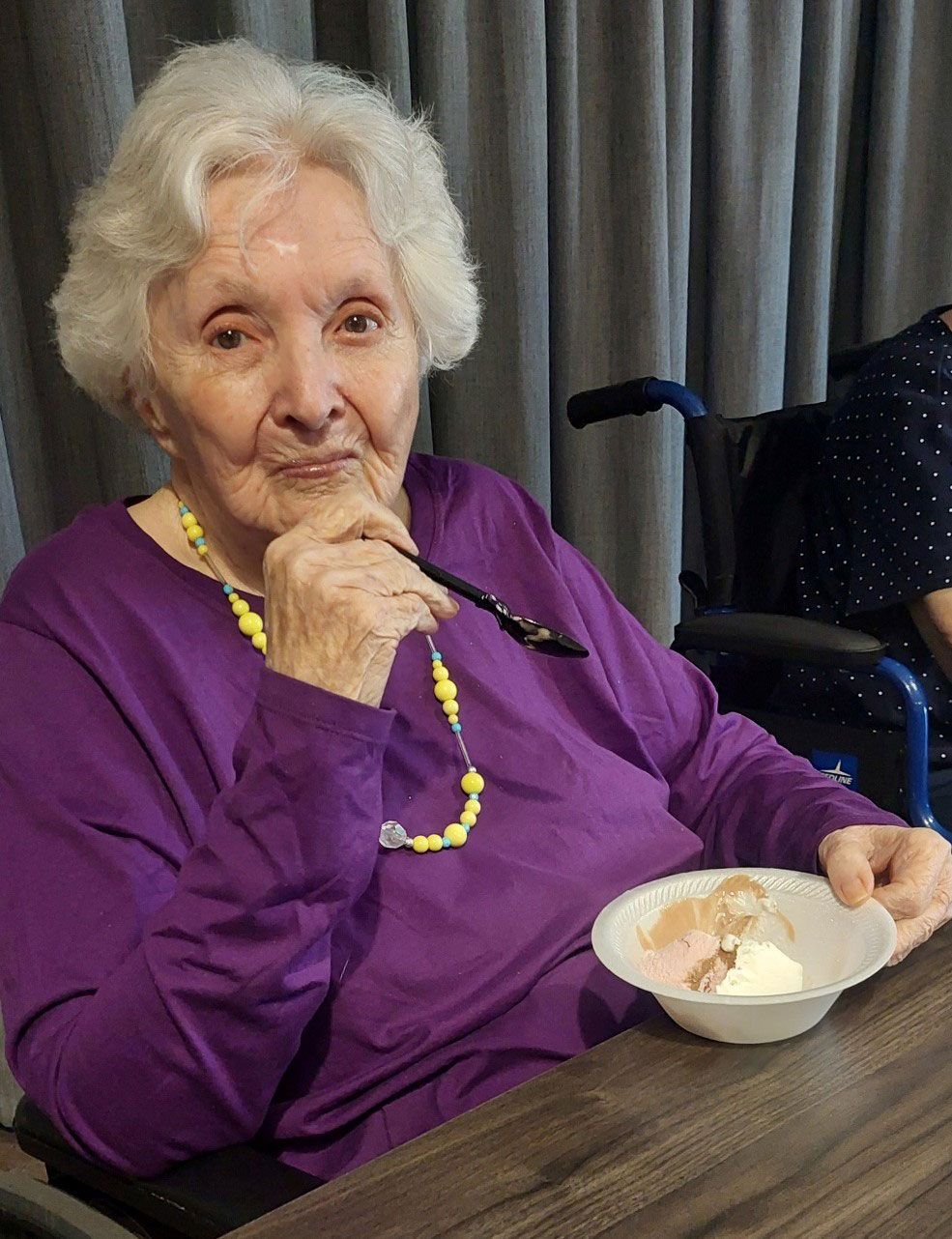 An individual in a purple shirt sits at a table, holding a spoon and a bowl of ice cream in a room with curtains.