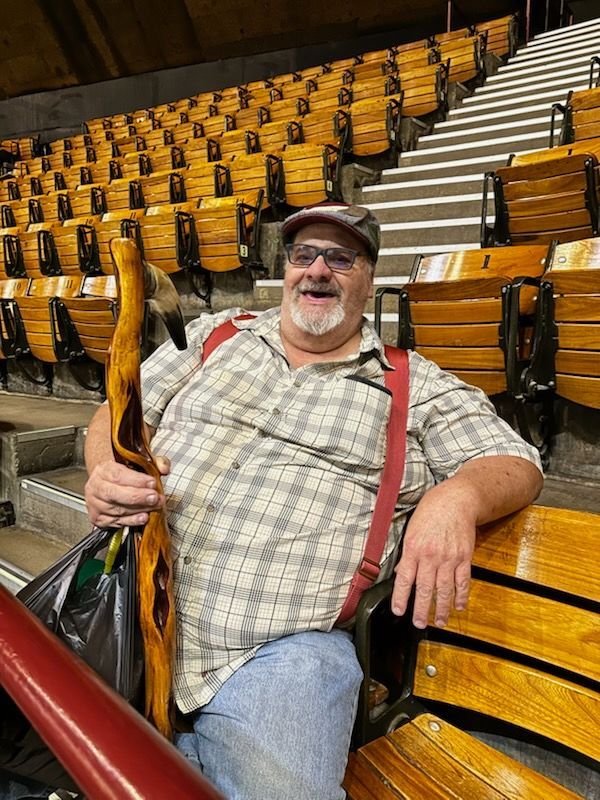 A person sitting in a stadium seat, smiling and holding a decorative wooden walking stick with red suspenders.