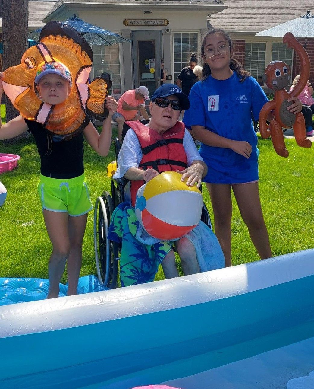 A person in a wheelchair wearing a life vest holds a beach ball between two children standing by an inflatable pool.