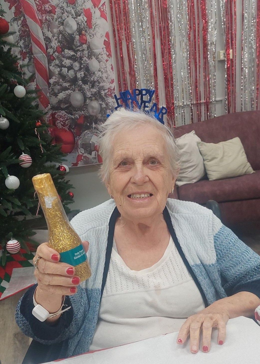 Smiling person in a blue cardigan holding a small gold glittery bottle, with a holiday-decorated tree in the background.