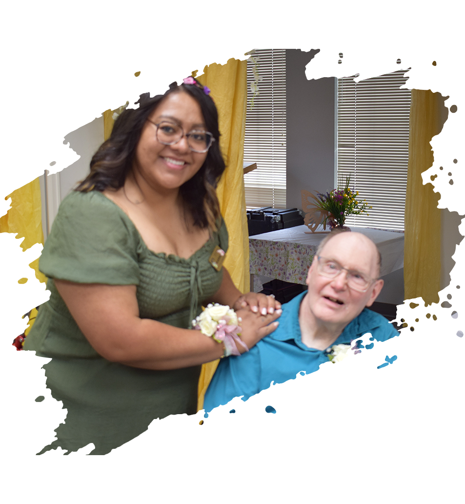 A person wearing a green top and a corsage on their wrist smiles while resting their hand on a person in a blue shirt.
