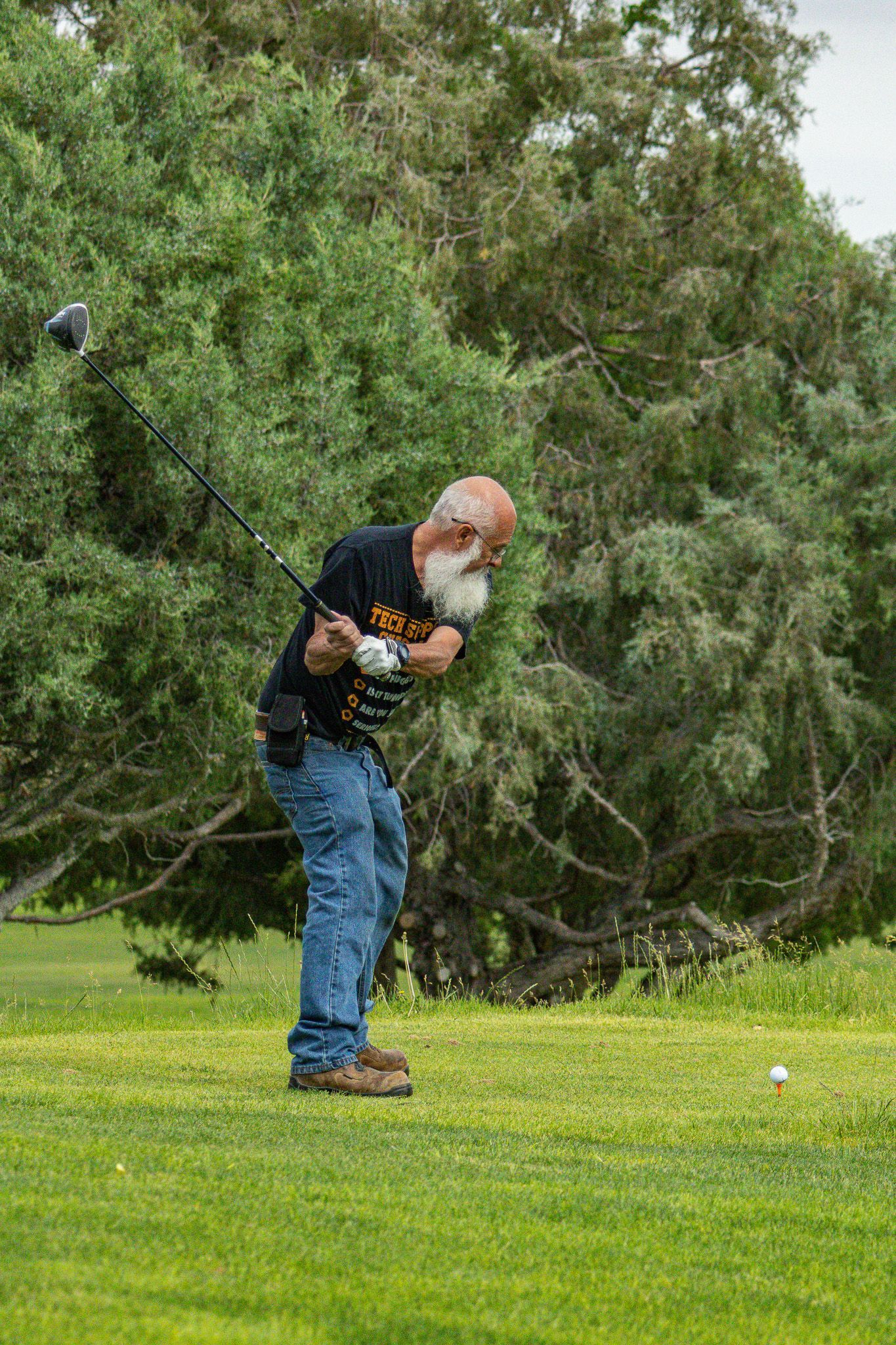 Golfer in blue jeans and black shirt mid-swing on a grassy course with trees behind him