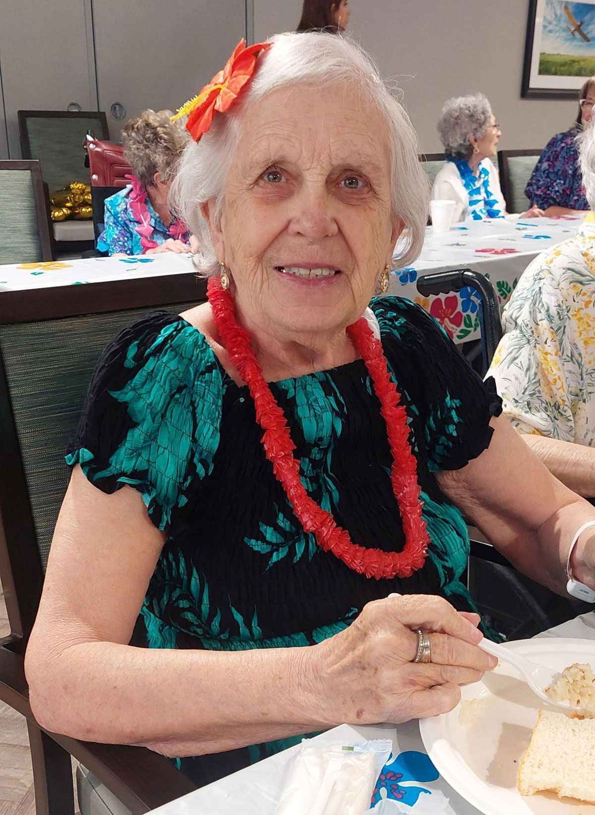 An smiling person wears a bright orange flower hair accessory and a red lei, seated at a table with food.