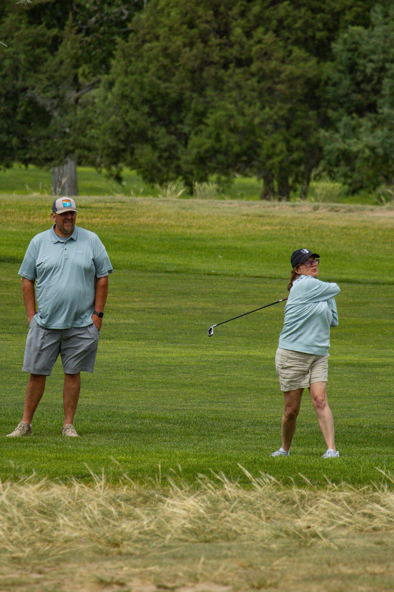 Two golfers on a green, one preparing to swing, with trees in the background.