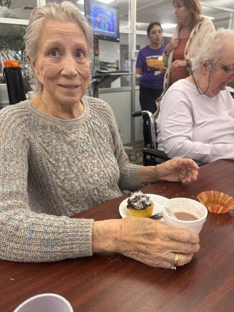 A person sitting at a table with a muffin and a drink, with others in the background in a room with glass partitions.