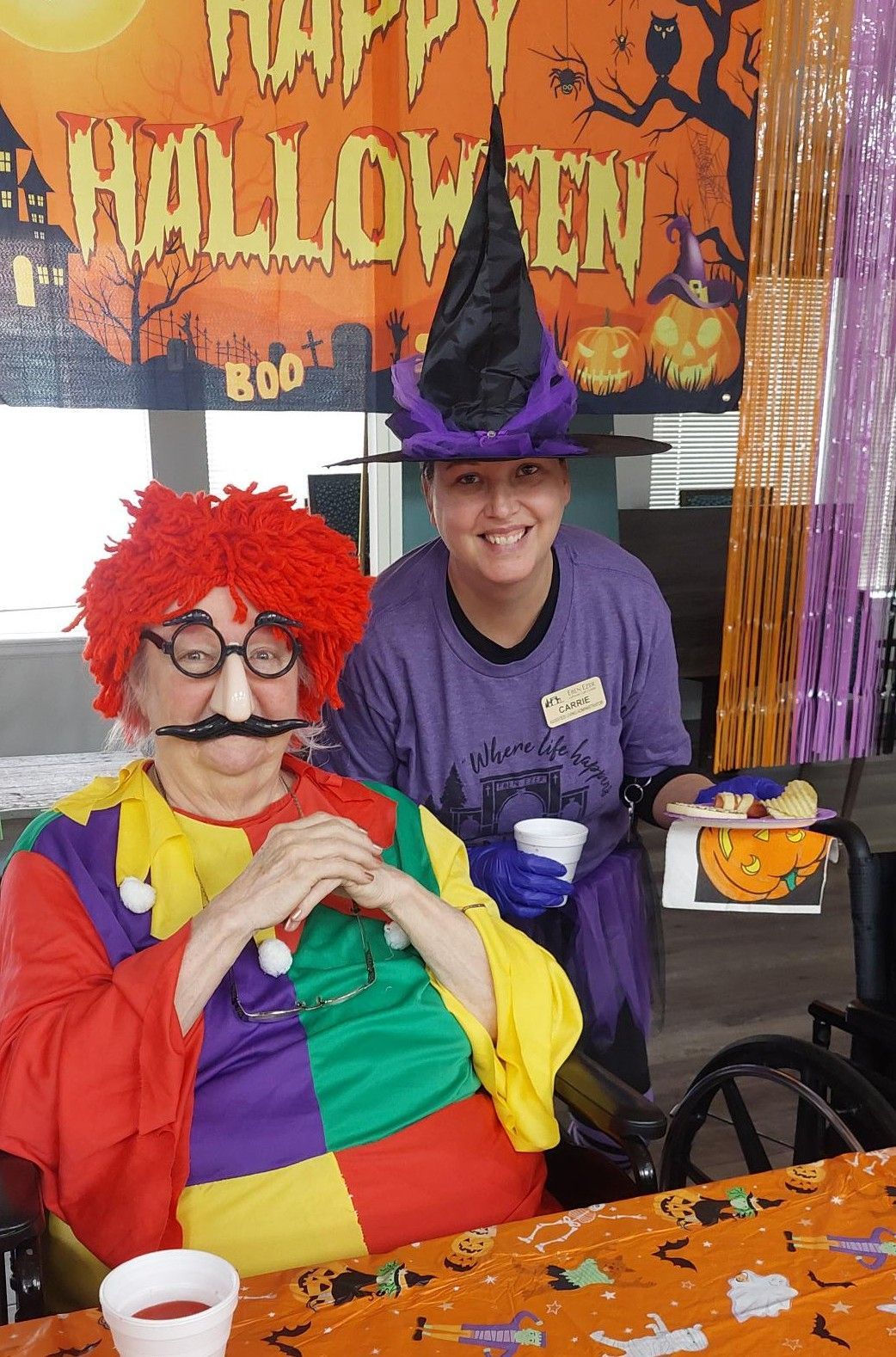 A person in a red wig and clown costume sits at a table near a person in a purple shirt and witch hat during Halloween.