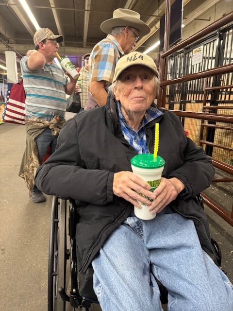A person in a wheelchair holds a cup with a straw in a building with metal stalls; others stand in the background.