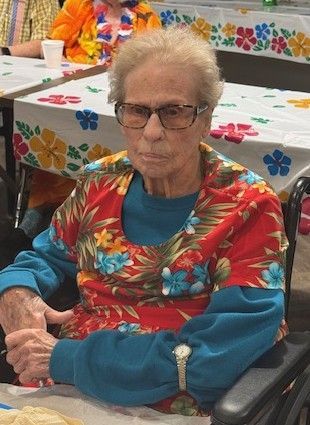 A person sits at a table with a floral tablecloth, wearing a teal long-sleeve shirt under a red floral vest and glasses.