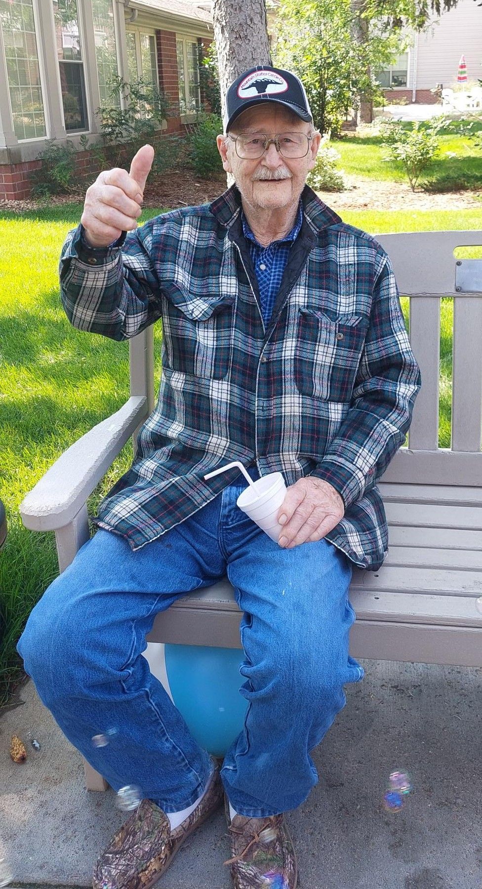 An elderly person wearing a plaid jacket, jeans, and a cap sits on a bench giving a thumbs-up while holding a white cup.