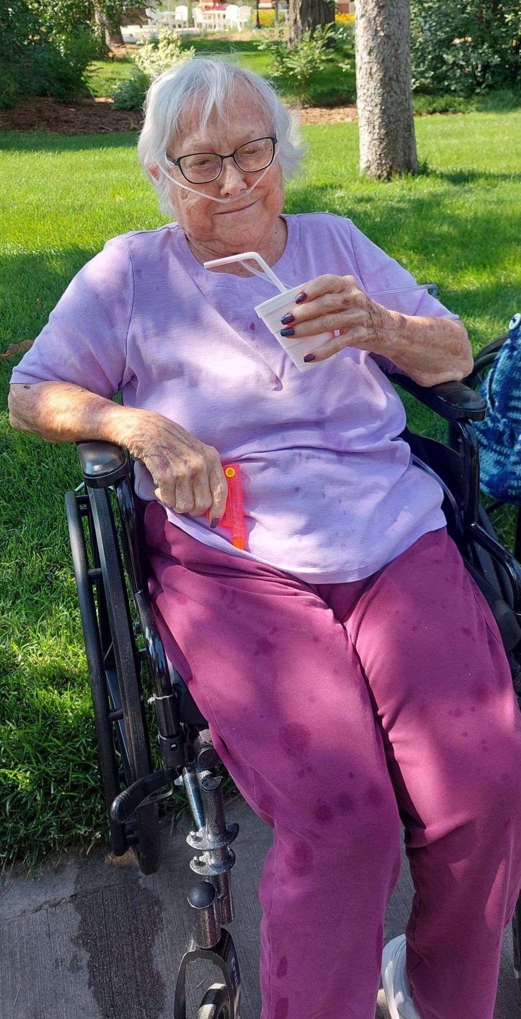 A person in a wheelchair, wearing a lavender shirt and purple pants, smiles while holding a drink in an outdoor setting.