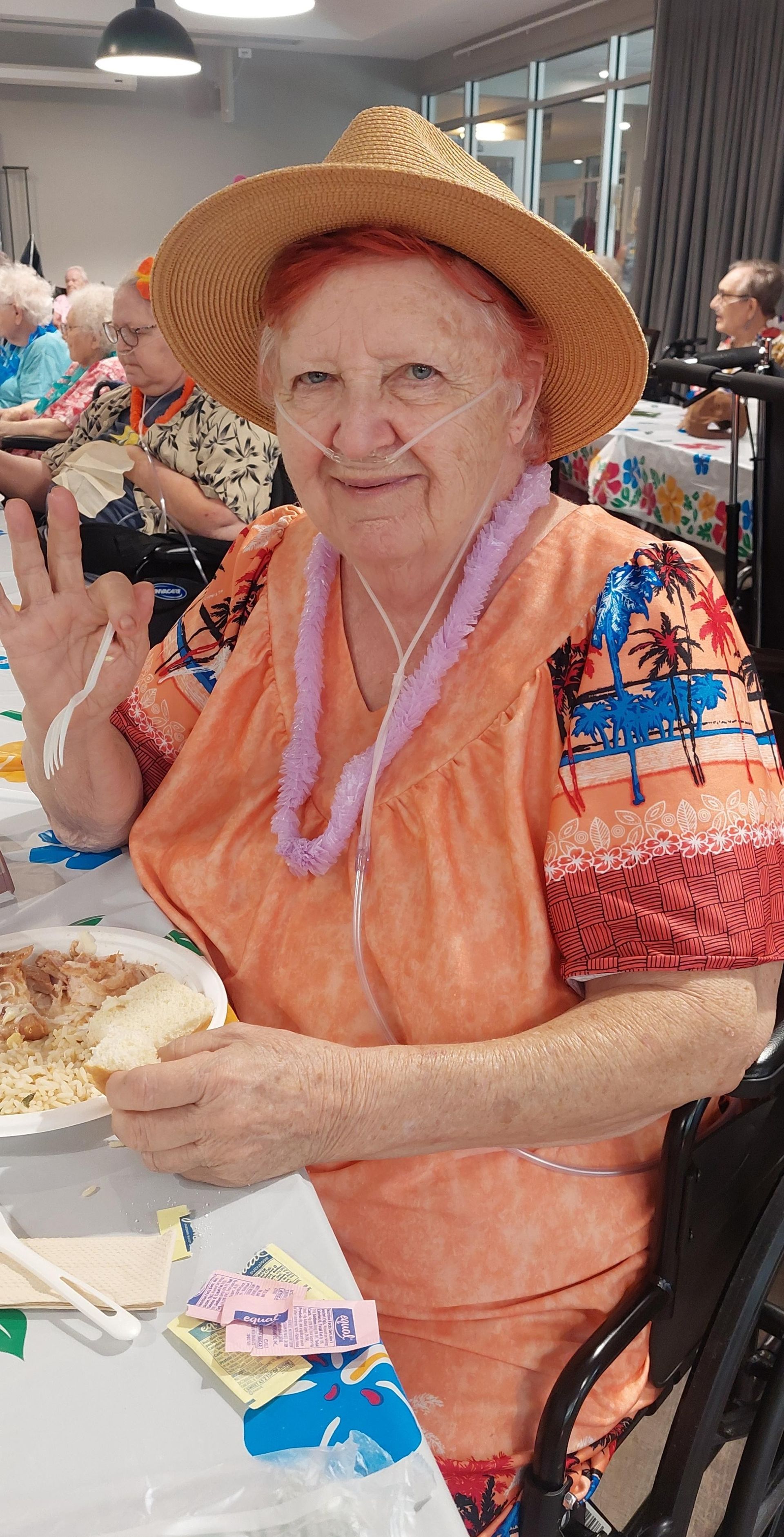 A person wearing a straw hat and orange patterned top with a lavender lei, smiling and making an OK gesture while dining.
