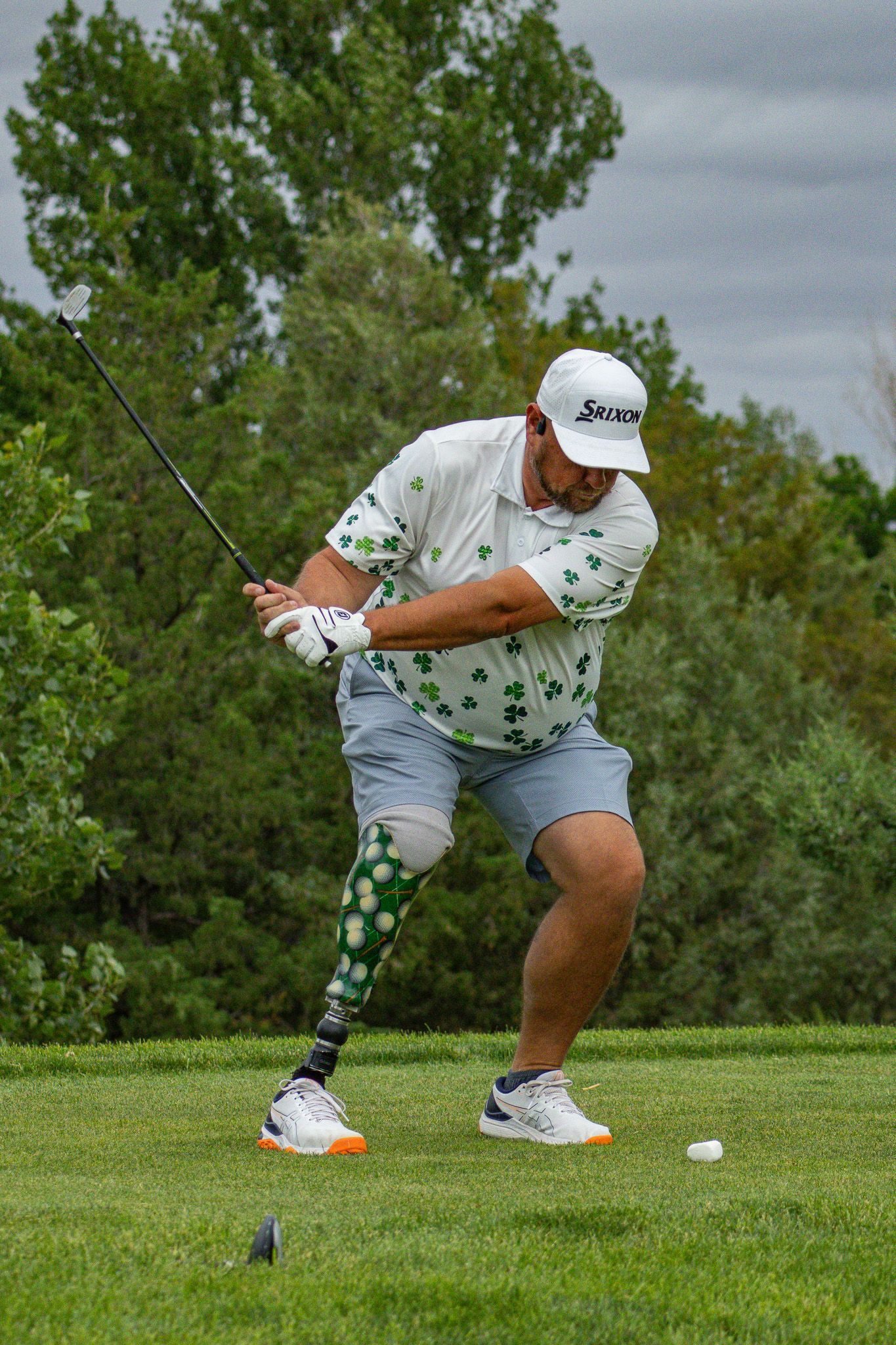 Golfer mid-swing on a grassy course, wearing a white cap and shirt, with trees in the background