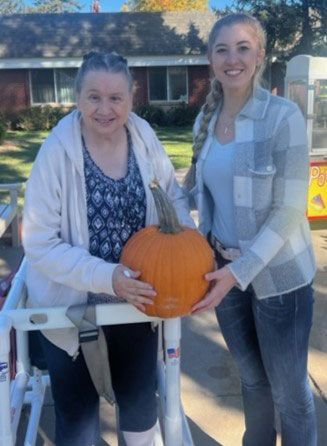 Two people smiling while holding a large orange pumpkin outdoors on a sunny day.