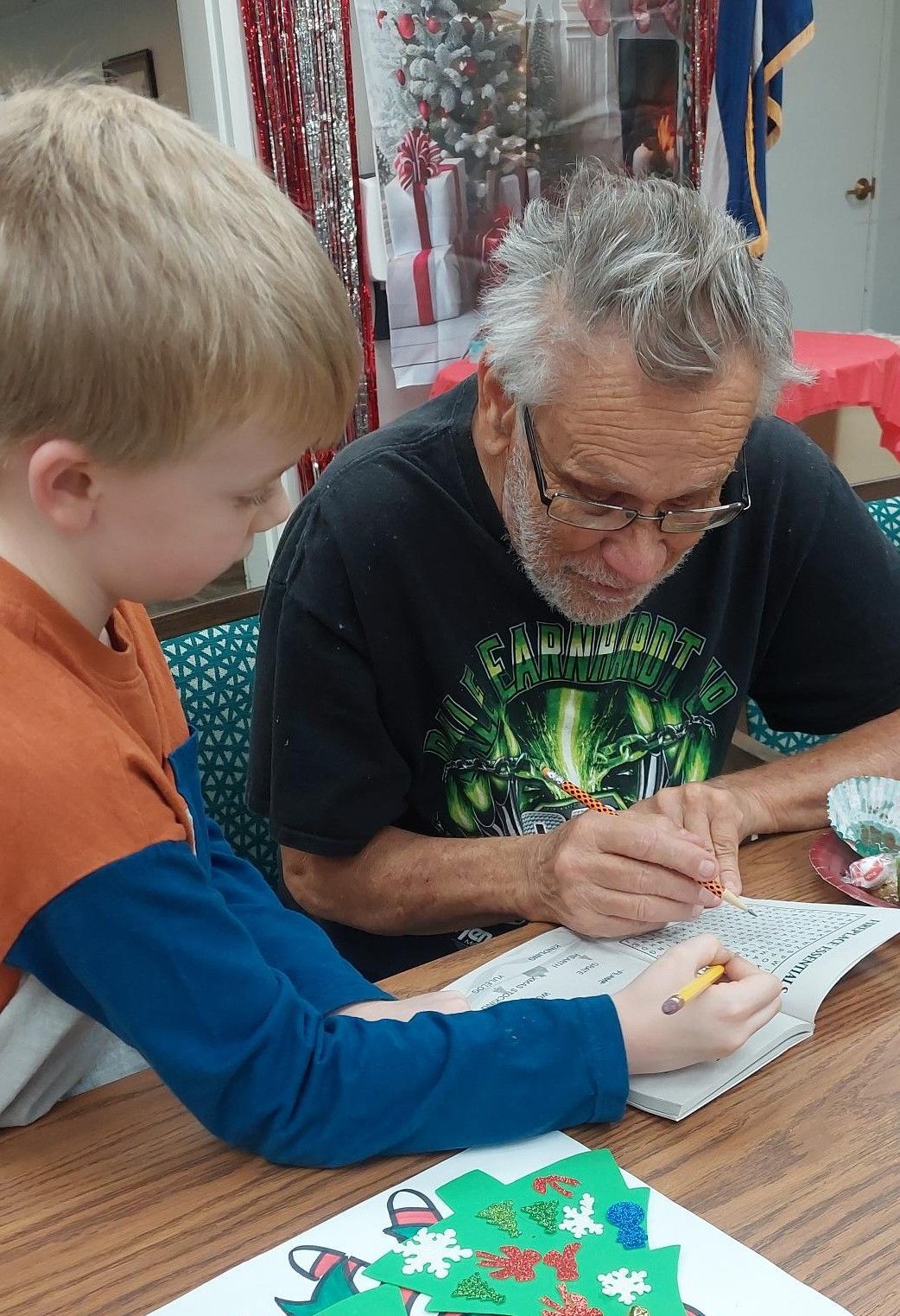 An adult and a child sit at a table together, both writing in a notebook near a Christmas-themed craft.
