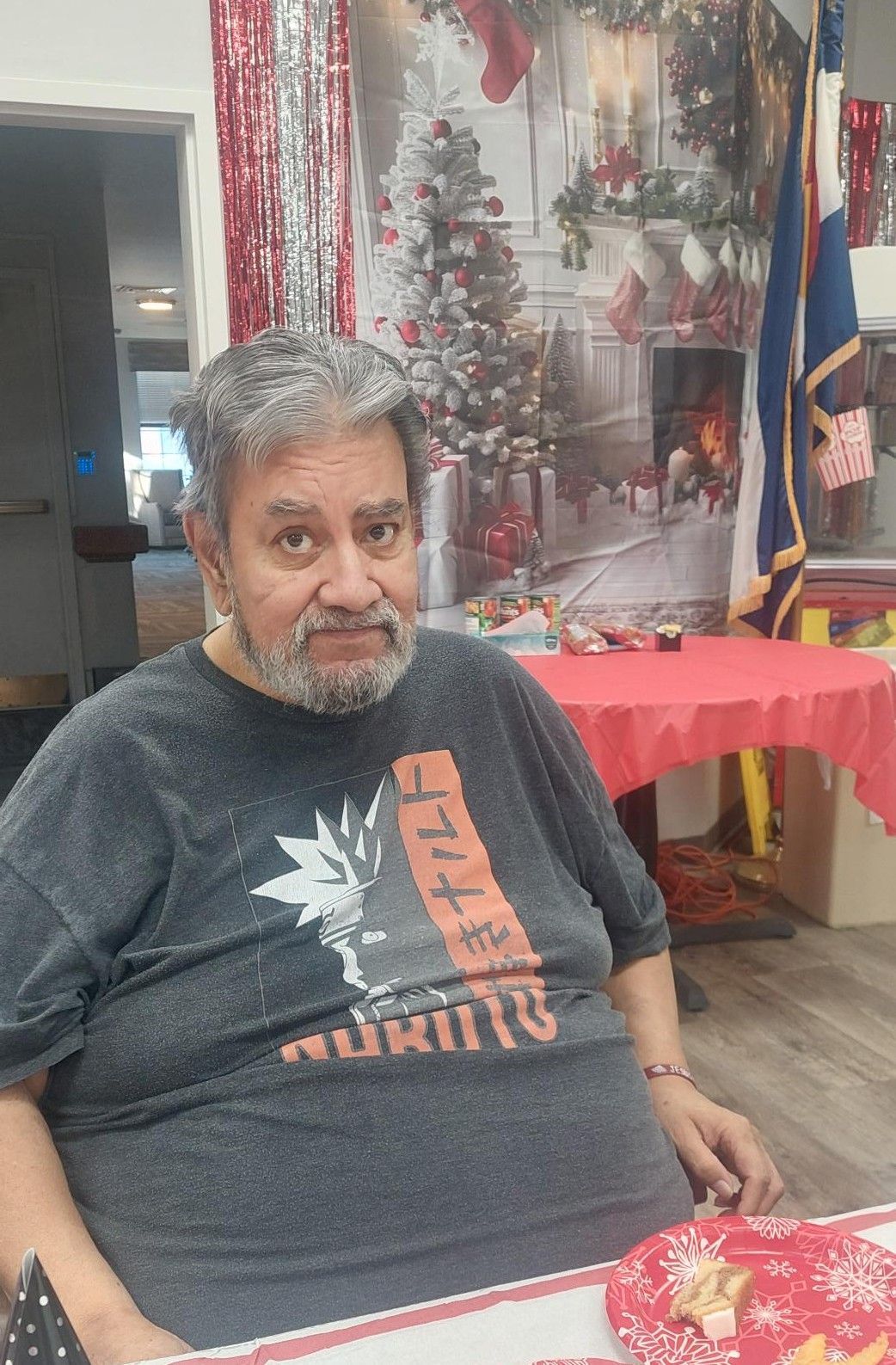 A person with a beard wearing a grey Naruto t-shirt sits at a table in a room decorated for a Christmas party.