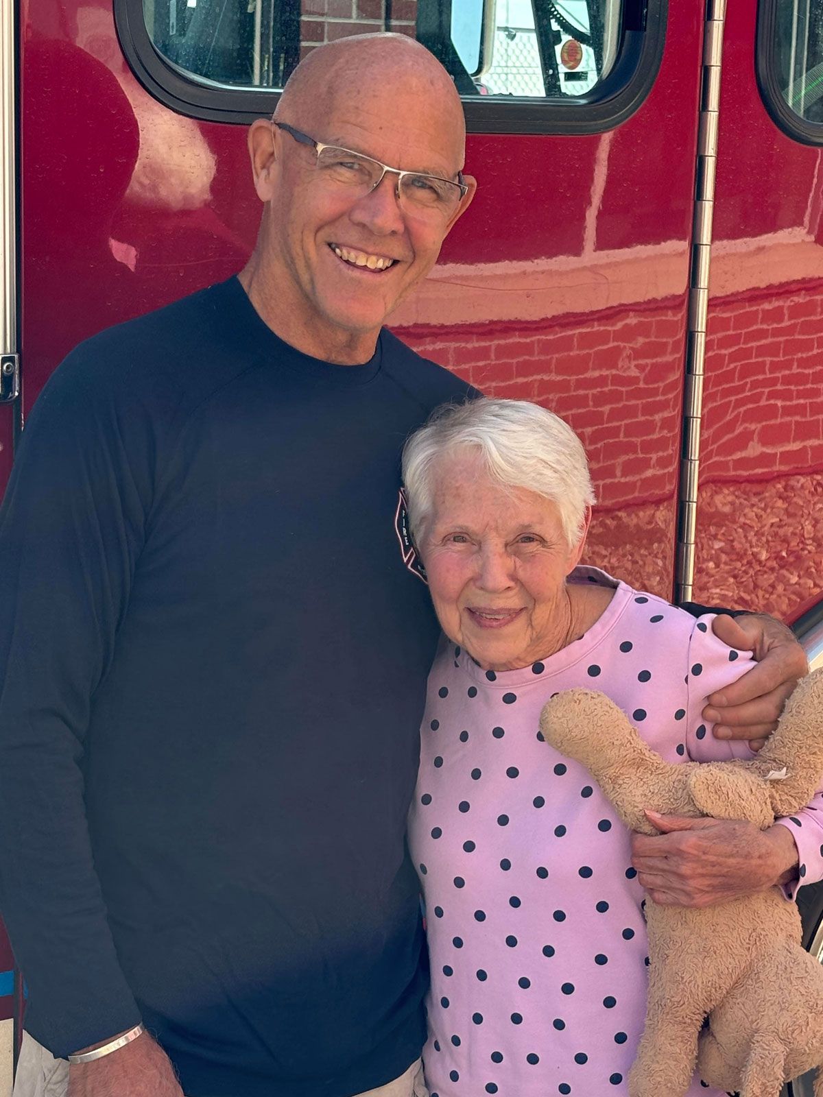 A smiling man in a dark shirt stands with his arm around a smiling person in a pink polka-dot top holding a toy animal.