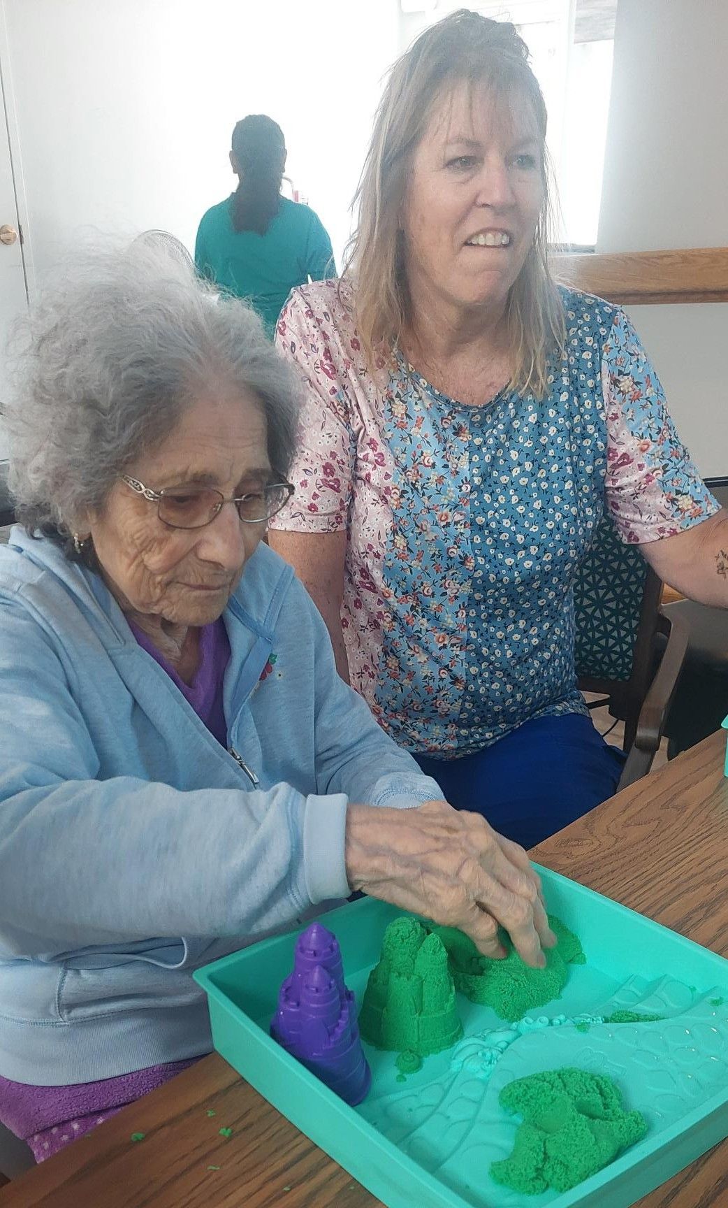 An older person and a caregiver interact while playing with green and purple kinetic sand in a tray on a wooden table.
