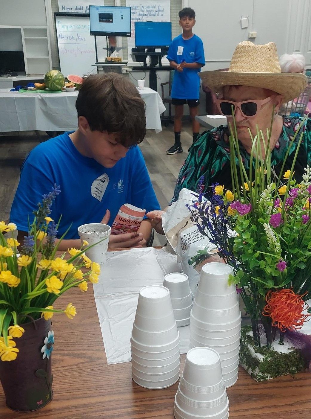 A person in a blue shirt works with someone wearing a straw hat and sunglasses at a table with white cups and flowers.