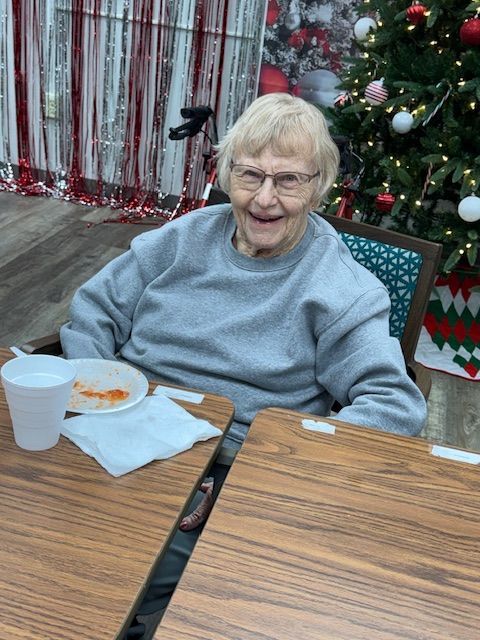 A person smiling while seated at a wooden table in front of a decorated Christmas tree and festive backdrop.