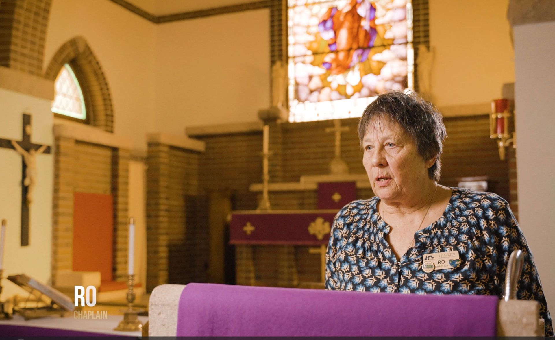 A person stands at a lectern with a purple cloth in a church, speaking with a serious expression before a stained glass.