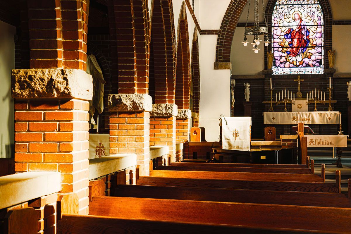 A view of a church interior with wooden pews, brick arches, and a stained glass window above the altar.