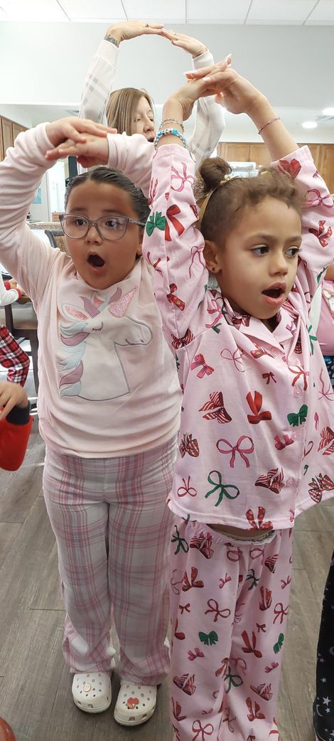 Two children in pajamas with arms raised above their heads, standing in an indoor setting with joyful expressions.