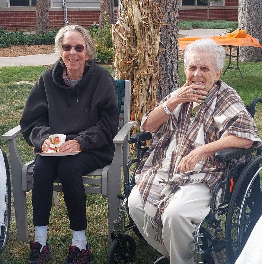Two people sitting outdoors, one in a chair holding a snack and the other in a wheelchair with a patterned shawl.