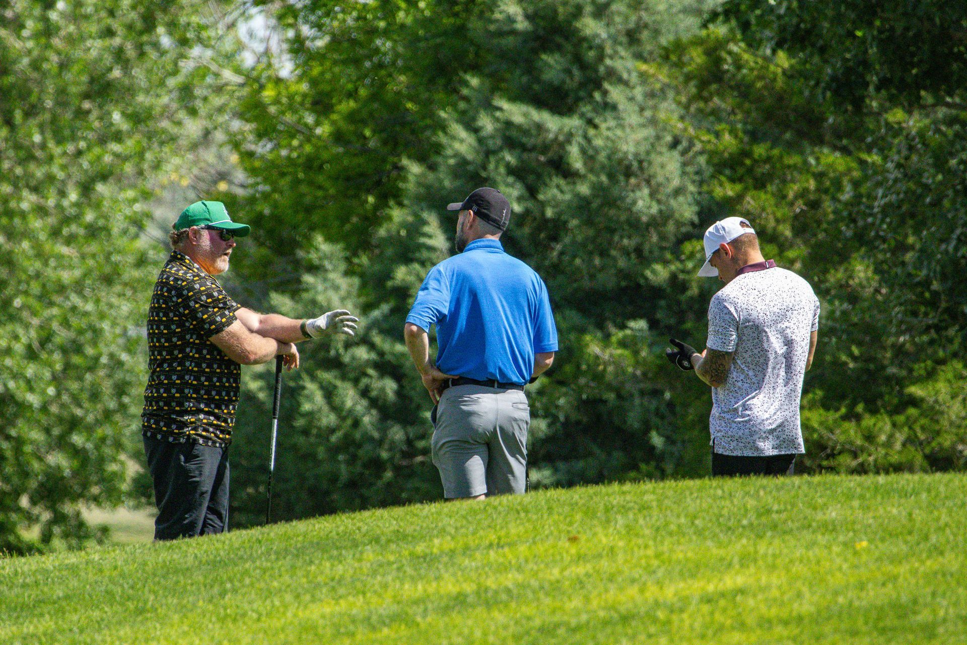 Three golfers standing on a green fairway, one holding a club and talking with the others.