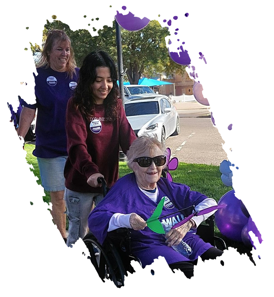 Two women planting a purple flower in a raised garden bed outdoors.