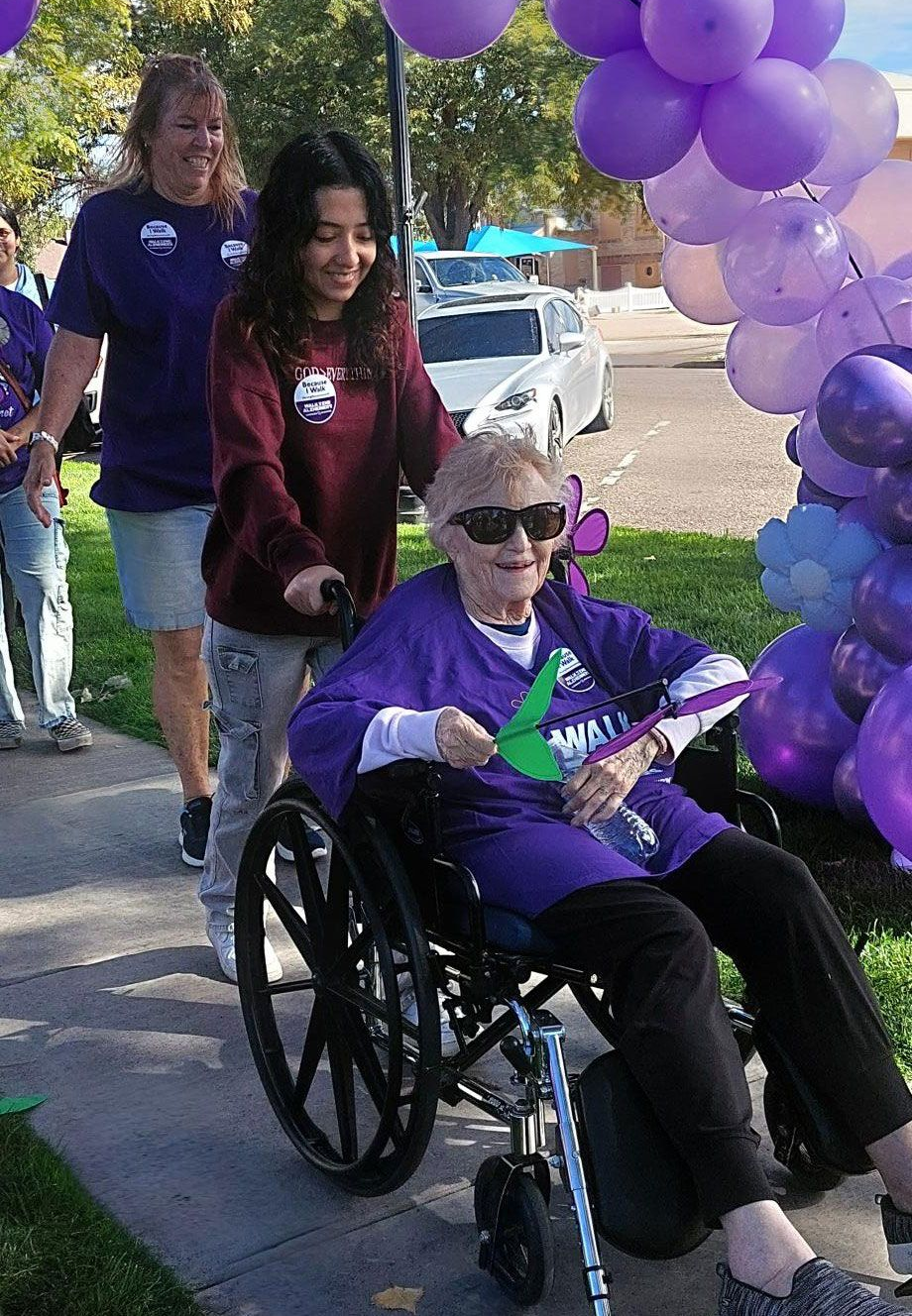 A person in a wheelchair wearing sunglasses and purple, accompanied by two people on a path near purple balloons.