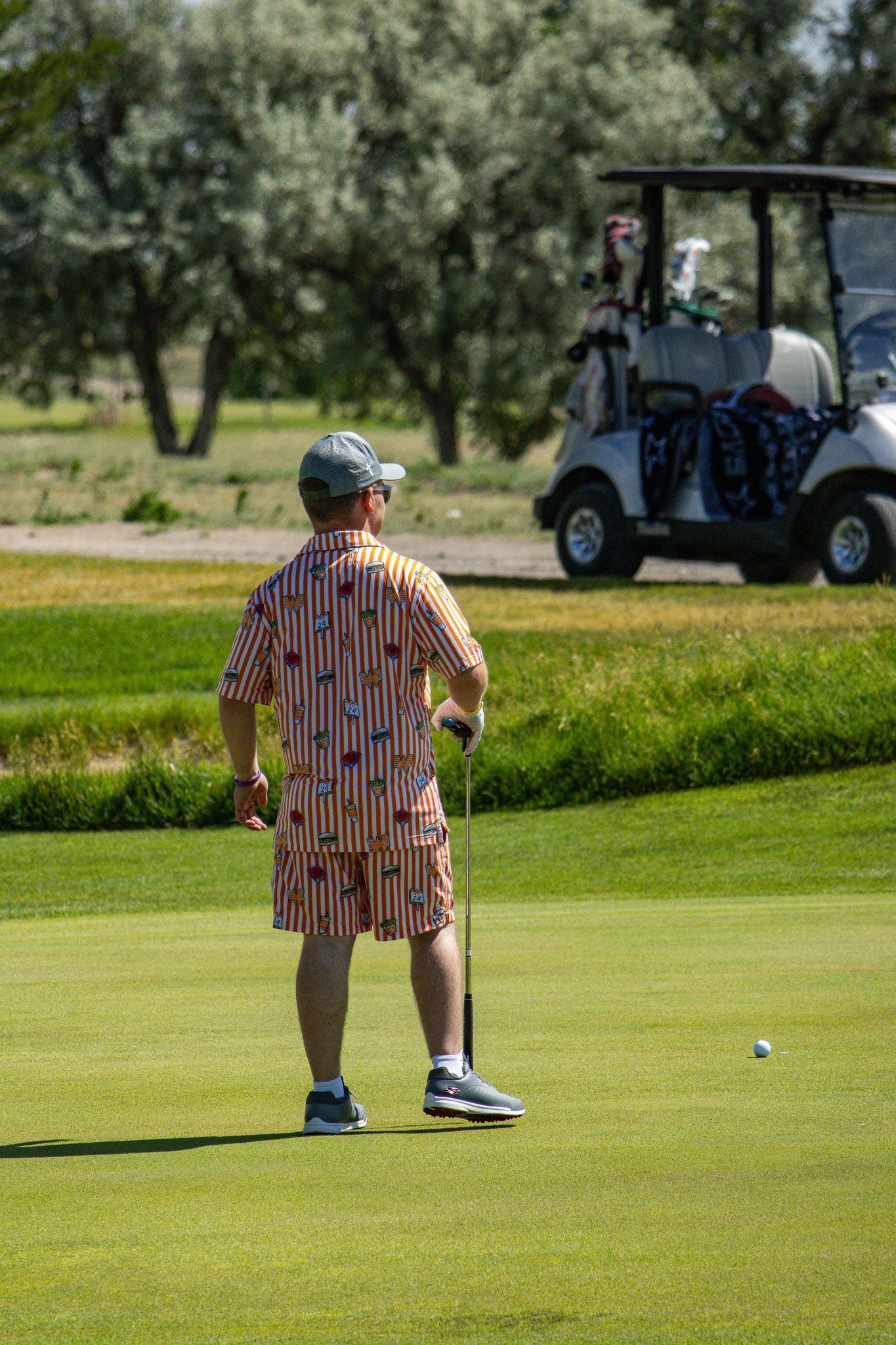 Golfer standing on green near a golf cart, with trees in the background.