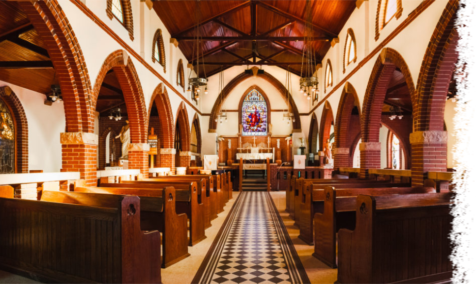 The interior of a church with wooden pews on both sides, a checkered tiled aisle, and arched brick supports.